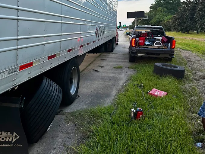 Commercial semi-trailer with a damaged tire on a highway shoulder while a service pickup and equipment are staged on the grass verge